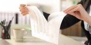 Mãos de uma pessoa em traje formal rasgando um documento de papel ao meio sobre uma mesa de escritório.